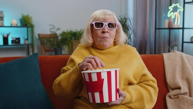 Senior old woman in 3D glasses on couch eating popcorn snacks and watching interesting TV serial sport game film online social media movie content at home apartment. Grandmother enjoying entertainment
