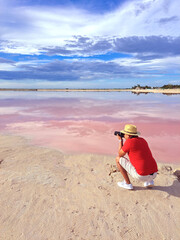 Latin adult photographer man with shorts, red shirt and hat takes pictures on the sand next to the pink colored lagoon with a high concentration of salt, Las Coloradas in Yucatan Mexico