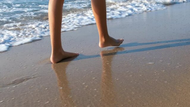 Teen bare legs in shore waves. A view of girl legs with bare foot walk along the wet beach by foaming sea in the sun light.