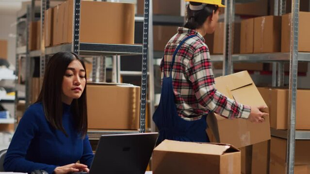 Young adult analyzing packs of goods in warehouse to help entrepreneur with distribution and business management. Asian team of people working on stock logistics shipment. Handheld shot.