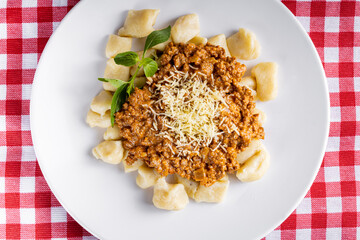 GNOCCHI COOKED WITH GROUND MEAT IN TOMATO SAUCE, COVERED IN GRATED CHEESE MOUNTED ON A WHITE PLATE ON A CHEERED TOWEL.