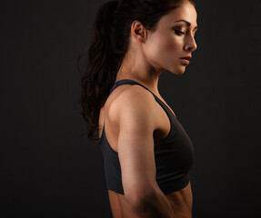 Fototapeta na wymiar Serious female sporty muscular with ponytail doing stretching workout the shoulders, blades and arms in sport bra, standing on dark shadow grey background with empty copy space. Profile