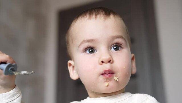 Grey-eyed Caucasian baby boy eating slowly from spoon. Some pieces of food falling from kid’s cheeks. Low angle view close up portrait.
