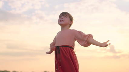 Little girl prays against background of sky and sun. Religion and god, childhood dreams. Happy family. Child raised his hands to sky in park at sunset, true faith. Child plays in park against sky.