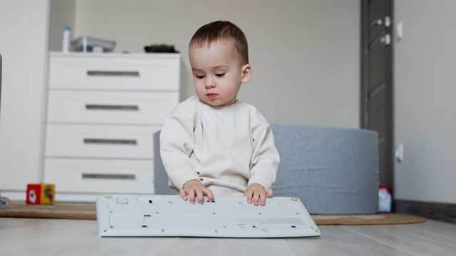 Calm baby boy sits on the floor playing with a computer keyboard. Kid stands up and walks away.