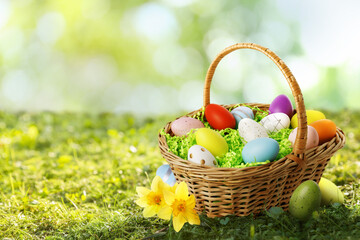 Wicker basket with festively decorated Easter eggs and daffodils on sunlit green grass, space for text