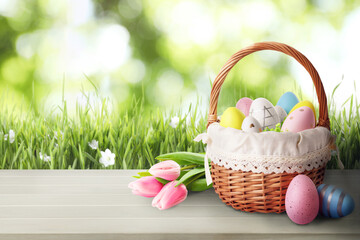 Fototapeta na wymiar Wicker basket with festively decorated Easter eggs and tulips on wooden table on green meadow, space for text