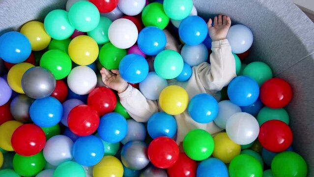 Baby boy lies hiding in the colorful balls in a dry pool. Toddler turns and sits in the basin taking one red ball in hands. Top view.