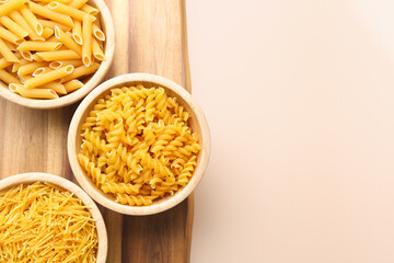 Wooden board with bowls of raw pasta on color background, closeup