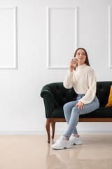 Young woman with cup of coffee sitting on green sofa near light wall