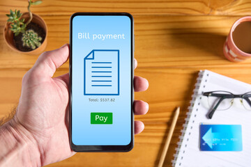 Close up of hand holding mobile phone with online bill payment app interface on the screen and green Pay button. Credit card and accessories on wooden table in the background - Powered by Adobe