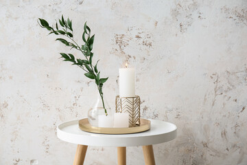 Burning candles and vase with ruscus branch on table near grey grunge wall