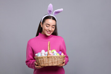Happy woman in bunny ears headband holding wicker basket of painted Easter eggs on grey background. Space for text