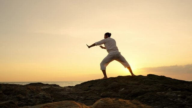 Caucasian man martial art practice and meditation with ocean nature on rocky coastal hill at summer sunset. Wellness people do outdoor relaxing yoga exercise. Health care and self motivation concept.