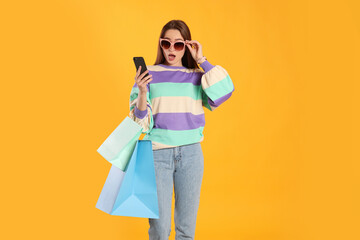 Surprised young woman with shopping bags looking at smartphone on yellow background. Big sale