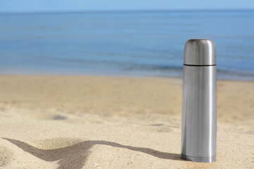 Metallic thermos with hot drink on sandy beach near sea, space for text
