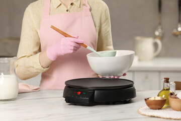 Woman cooking delicious crepe on electric pancake maker at white marble table in kitchen, closeup
