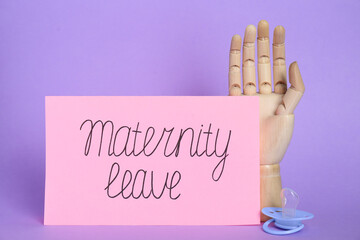 Wooden mannequin hand, card with phrase Maternity Leave and pacifier on violet background
