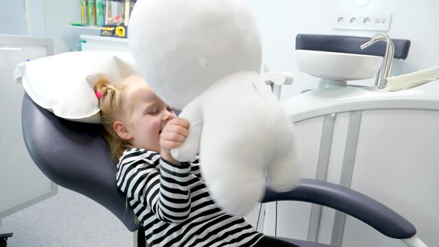 4.5-year-old girl plays in a dental chair with soft toy Lunatic Teletubby or an astronaut she waves it looks smiles relaxed waits for a doctor pediatric dentistry acquaintance protection care