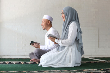 Side view of Asian muslim couple smiling while holding Quran on the prayer mat at the mosque. 