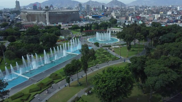 4k video of a water fountain park with many jets praying water upwards. Trees and grass surround the fountains. A stadium and cityscape in background. Drone flies forward. Located in Lima, Peru.