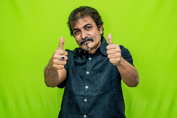 The well-dressed Indian man wearing a black shirt appears to be celebrating a win, as he raises both hands and gives a thumbs-up gesture while standing against a green screen background - Powered by Adobe