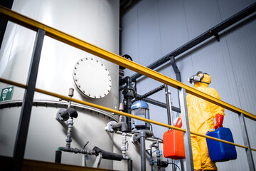 Worker supervisor in protective yellow suit, gas mask walking around large industrial acid tank and...