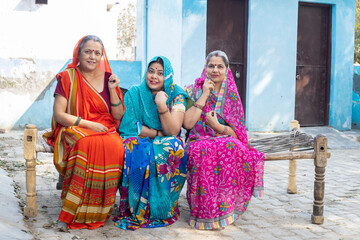 Rural indian women wearing colorful sari or saree sitting on wooden bed at village looking at camera.