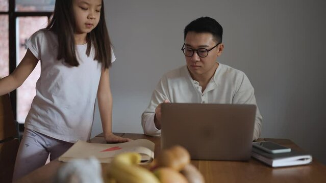 Confident Asian father works on his laptop while sitting at the table with his daughter in the kitchen