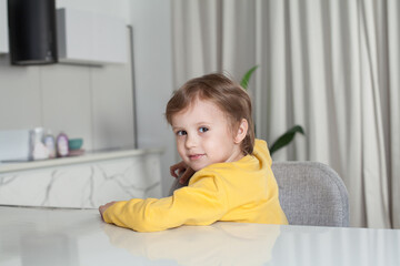 Pensive child boy with brown hair in yellow sweater sitting sideways by the table on studio kitchen background