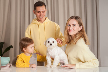 Happy adorable parents in yellow sweaters with child son and white dog, three abstract plastic bottles on the table