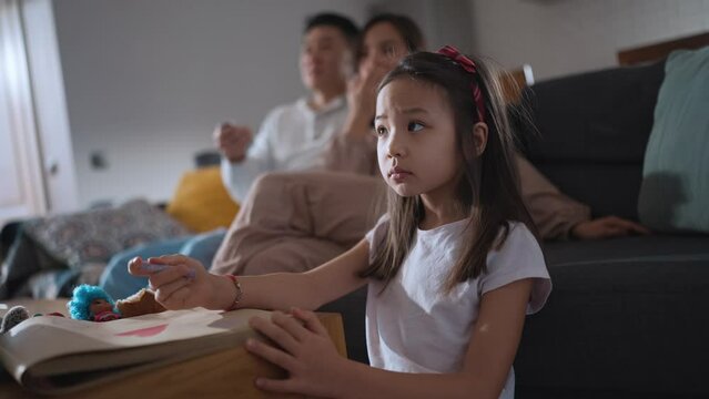Serious Asian young girl watching TV with her family and drawing at home