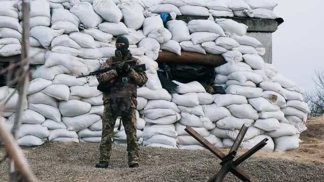 Soldier with weapon in military uniform stands in front of the barricades from sandbags and anti-tank hedgehog barriers. Military man on the roadblock. Combatant in full ammunition. Concept of war