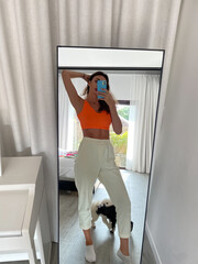 Fit tanned woman in orange top and cosy comfy pants at home take photo selfie on phone in mirror for social media, stories, vertical.