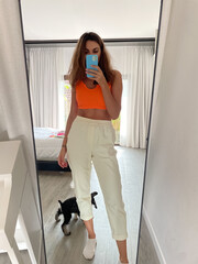 Fit tanned woman in orange top and cosy comfy pants at home take photo selfie on phone in mirror for social media, stories, vertical.