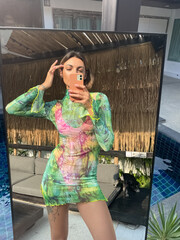 Fit sporty tanned woman in green short transparent dress outdoor at home take photo selfie on phone in mirror for social media, stories, vertical by the pool
