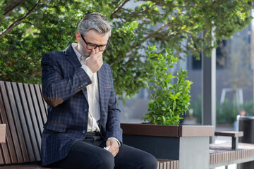 Sick and cold mature adult man sitting on bench outside office building, businessman in business suit sneezing and having runny nose seasonal allergy, employee boss during work break on bench.