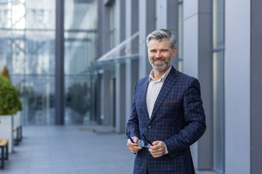 Portrait of happy successful smiling gray haired businessman, man in business suit standing outside office building smiling and looking at camera, satisfied mature boss investor holding glasses.