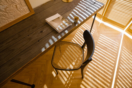 View from above on wooden table and chair on the floor made of parquet laid with herringbone. Shadows from the blinds on the windows. Fragment of the interior in beige tones in boho style