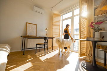Young woman stands with her dog by the window blinds and looks away in cozy and sunny living room of modern apartment in beige tones. Quarantine, loneliness and life at home concept - 578950189