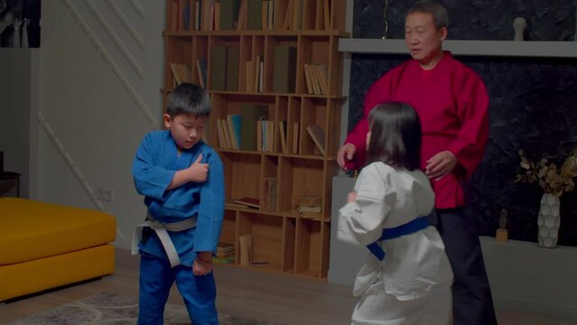 Senior handsome black belt taekwondo master teaching active motivated elementary age children in uniform to martial arts, explaining blocking techniques to avoid high kick during indoor training.
