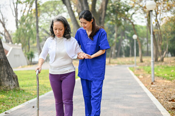 Caring Asian female caregiver and a senior lady with a walking stick are walking in the park