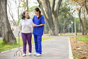 Smiling Asian female caregiver and a senior lady with a walking stick are walking in the park