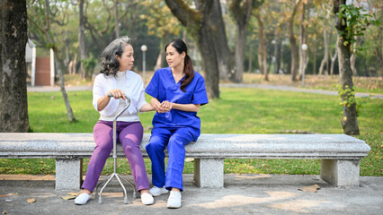 Fototapeta na wymiar Asian female caregiver is talking while sitting on a bench in a park with an old disabled lady.
