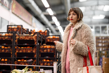 Woman buying fruits at local indoor market choosing mandarins during winter time. Girl in fur coat...