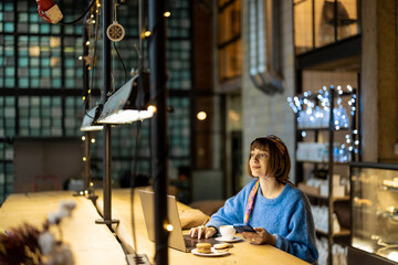 Young stylish woman works on laptop while sitting with a coffee drink at modern cafe. Concept of remote work from public place, digital freelance and modern lifestyle