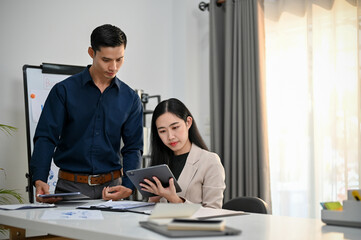 Fototapeta premium Two professional Asian businesspeople looking at tablet screen, discussing and brainstorming