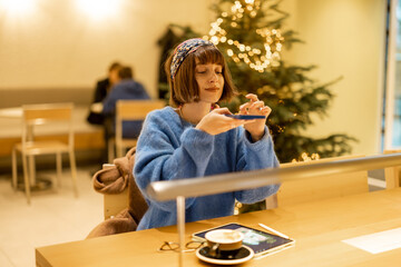 Young adult woman works on a digital tablet and phone while sitting at modern coffee shop with Christmas tree on background. Concept of remote creative work online