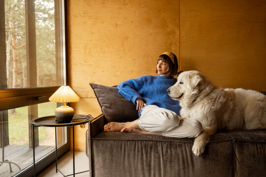 Portrait of a young woman with her dog sitting together on a couch and looking at window with view on the forest. Resting in countryside cottage and friendship with pets concept