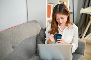 Charming Asian businesswoman working from home, using laptop and sipping coffee on sofa